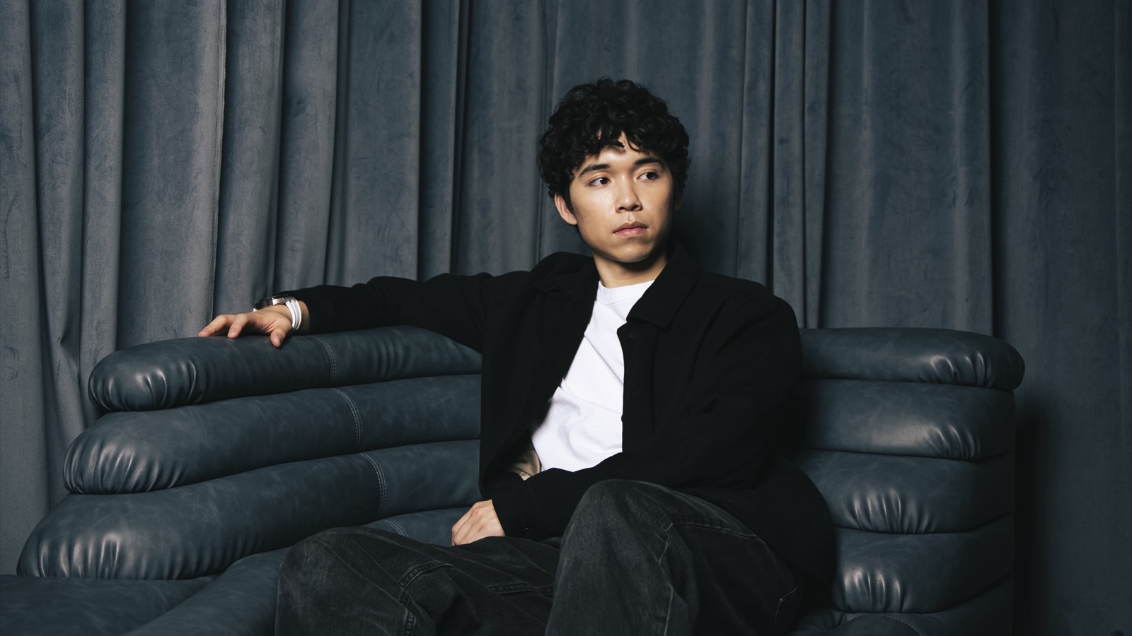 Asher Lara seated on a dark leather sofa in front of gray curtains