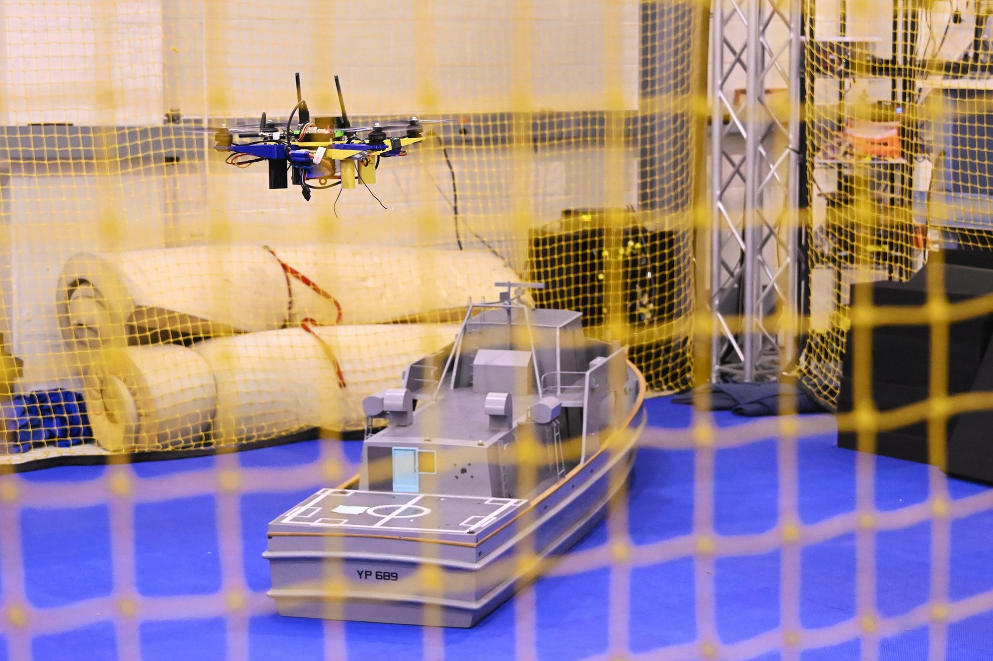 A small quadcopter hovering over a grey scale model of a navy patrol boat inside a netted test area
