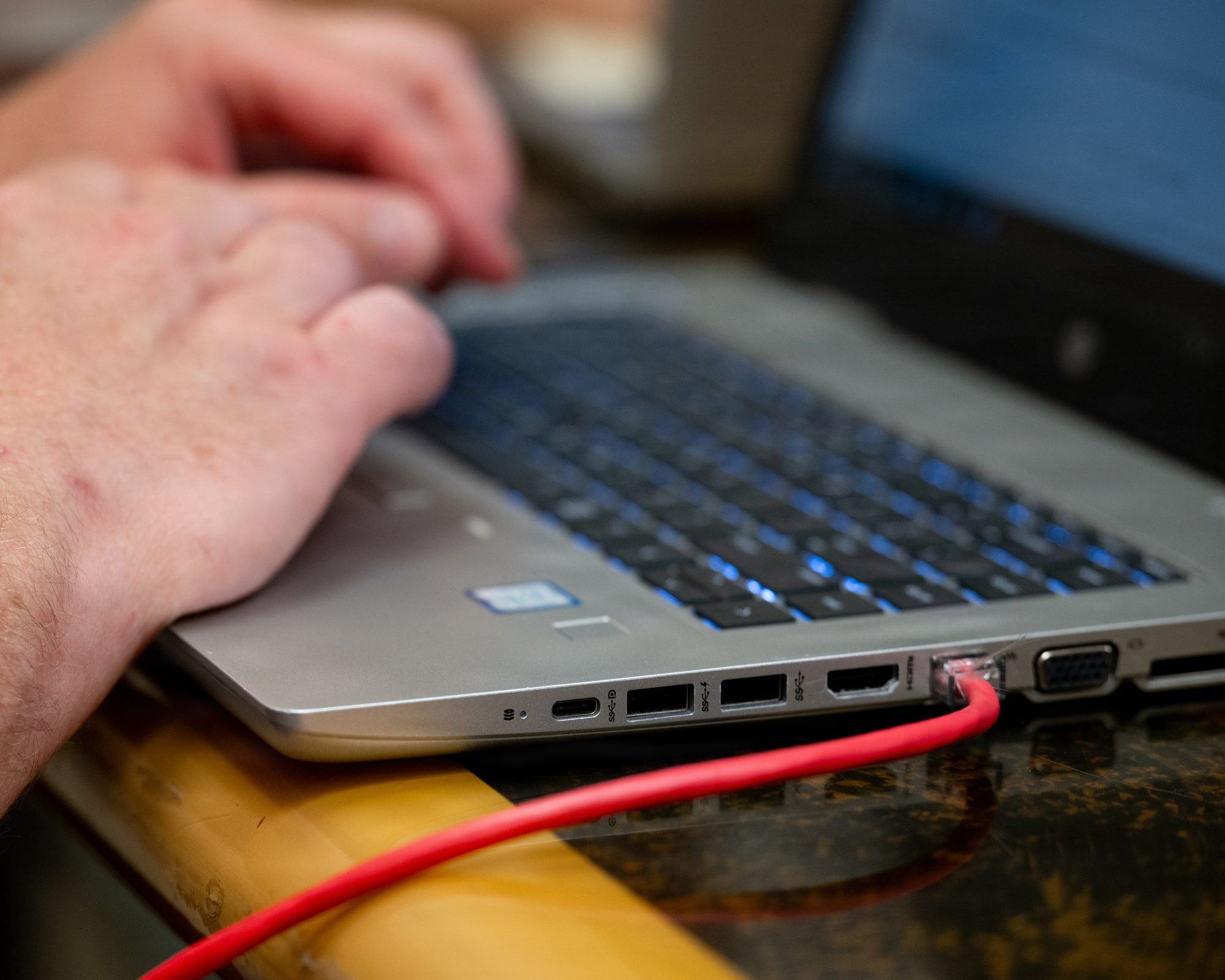 A hand resting on a silver laptop keyboard, with a red network cable plugged into the laptop's side