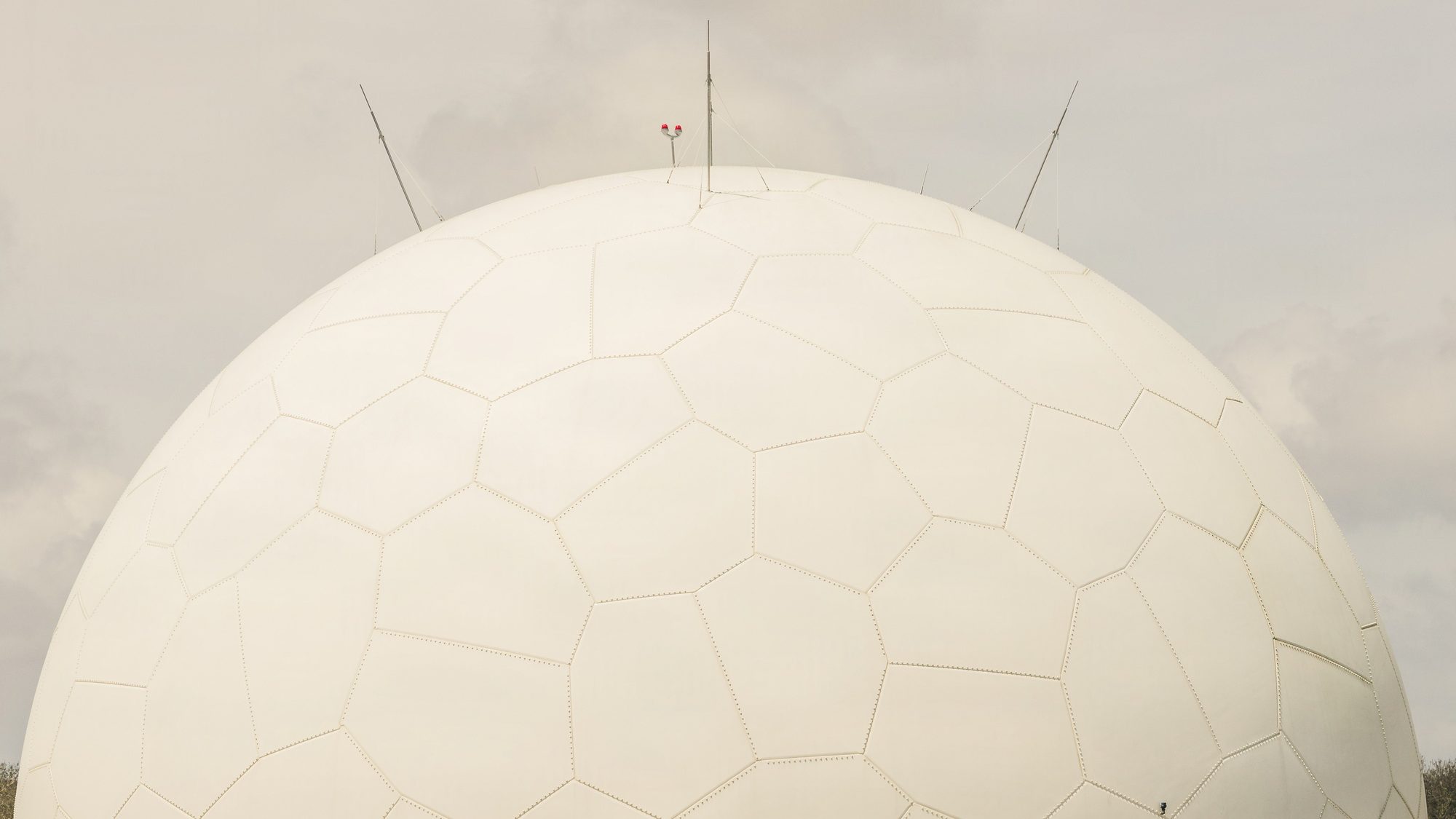 The top of a large white radar dome with antennas against a grey sky